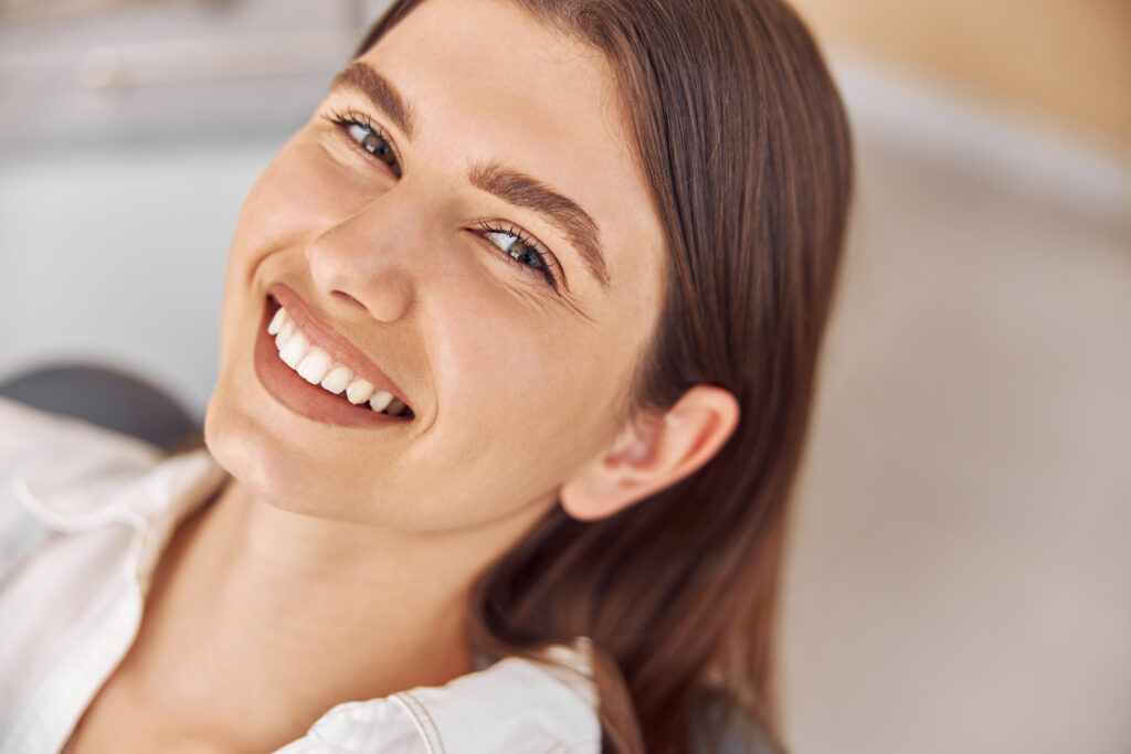 A close-up, slightly high-angle shot of a smiling woman with long brown hair, wearing a white collared shirt, showing off her teeth. The background is soft and brightly lit, suggesting an indoor setting like a dentist's office.