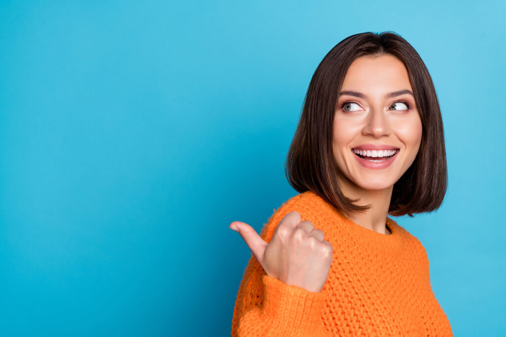 A cheerful young woman in an orange sweater smiling and pointing her thumb to the side against a blue background.
