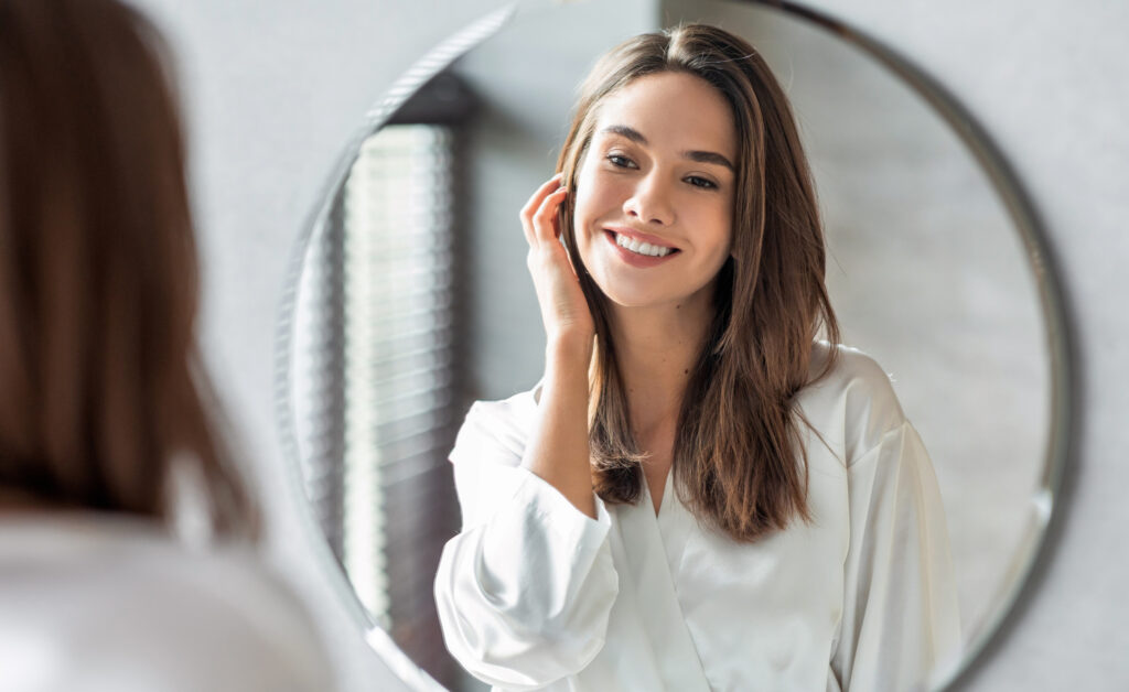 A smiling young woman with brown hair, wearing a white silk-like robe, looks at her reflection in a round bathroom mirror and gently touches her hair.