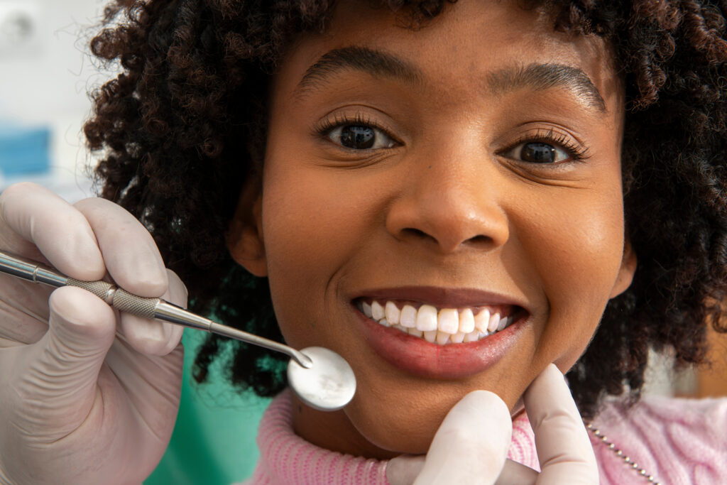 A close-up of a young woman smiling at the dentist, while gloved hands hold a dental mirror and gently touch her chin.