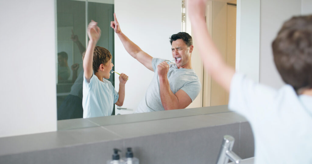 A man and a young child dance and sing energetically in front of a bathroom mirror, while the child holds a toothbrush.