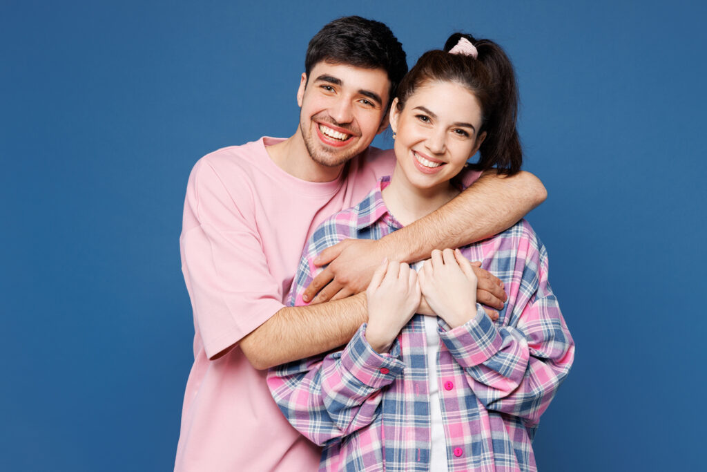 Young man in a pink shirt hugs a smiling young woman in a plaid shirt, both standing in front of a blue background.