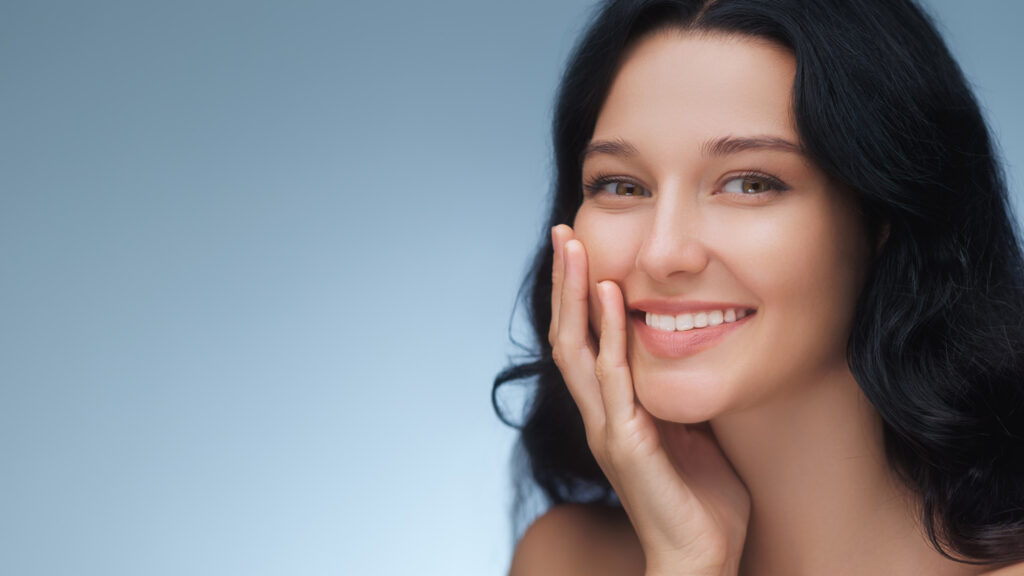 A portrait of a woman with dark wavy hair smiling and gently touching her cheek, showcasing her white teeth against a soft blue background.
