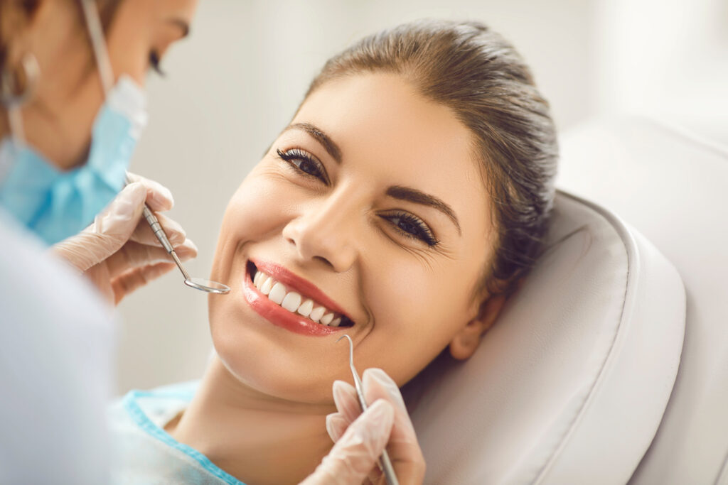 Close-up of a smiling young woman lying in a dental chair while a dentist in blue gloves examines her white teeth with metal tools.